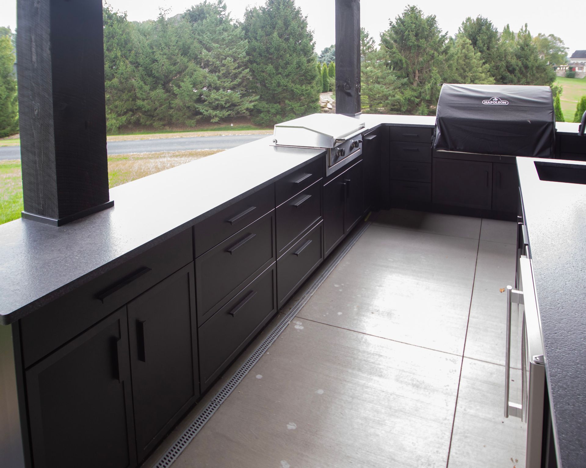 Covered outdoor grill station with black cabinets, countertop, and stainless grill by a grassy road.