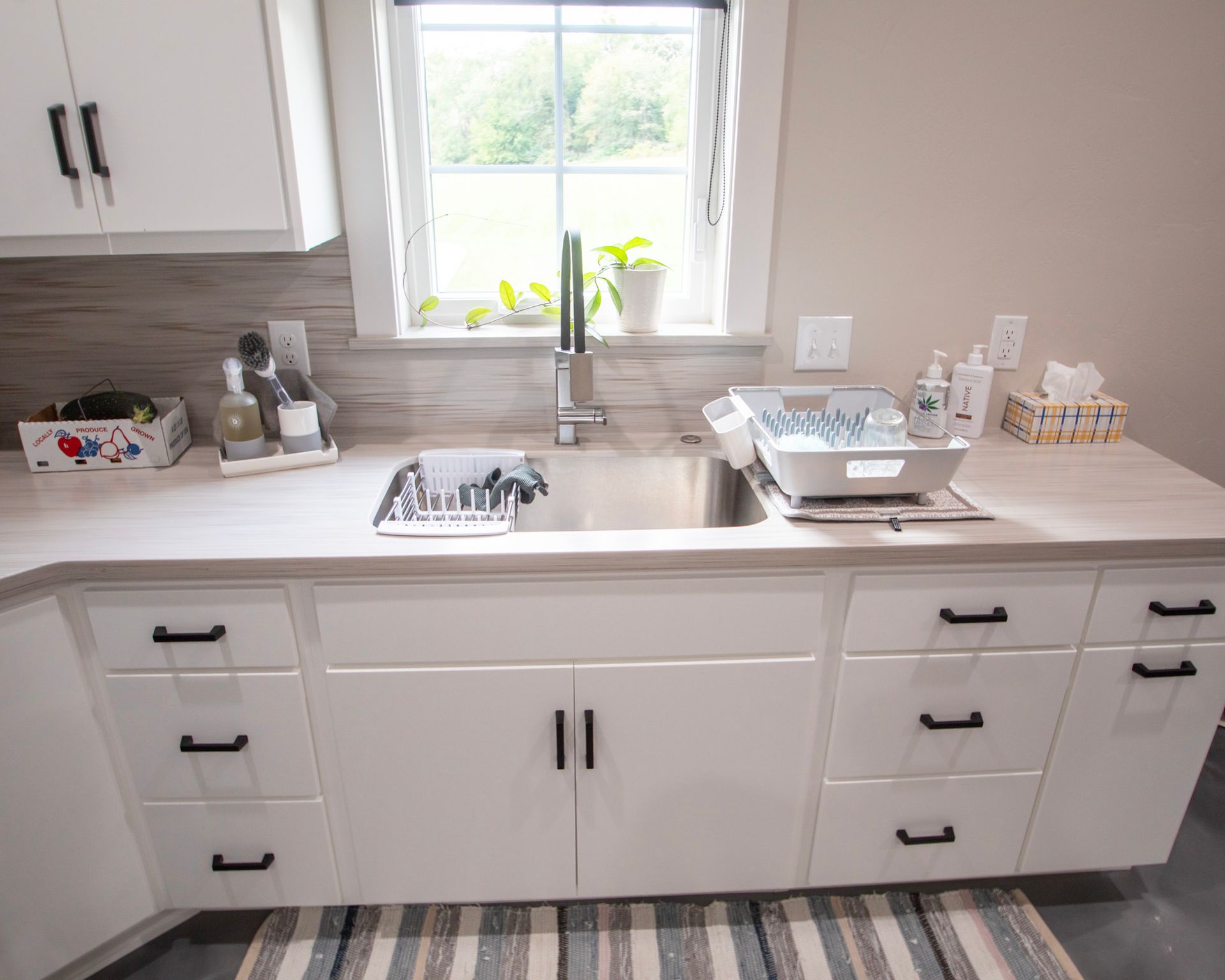 Bright white kitchen sink with window, cabinets, countertop trays, and dish rack on the right.