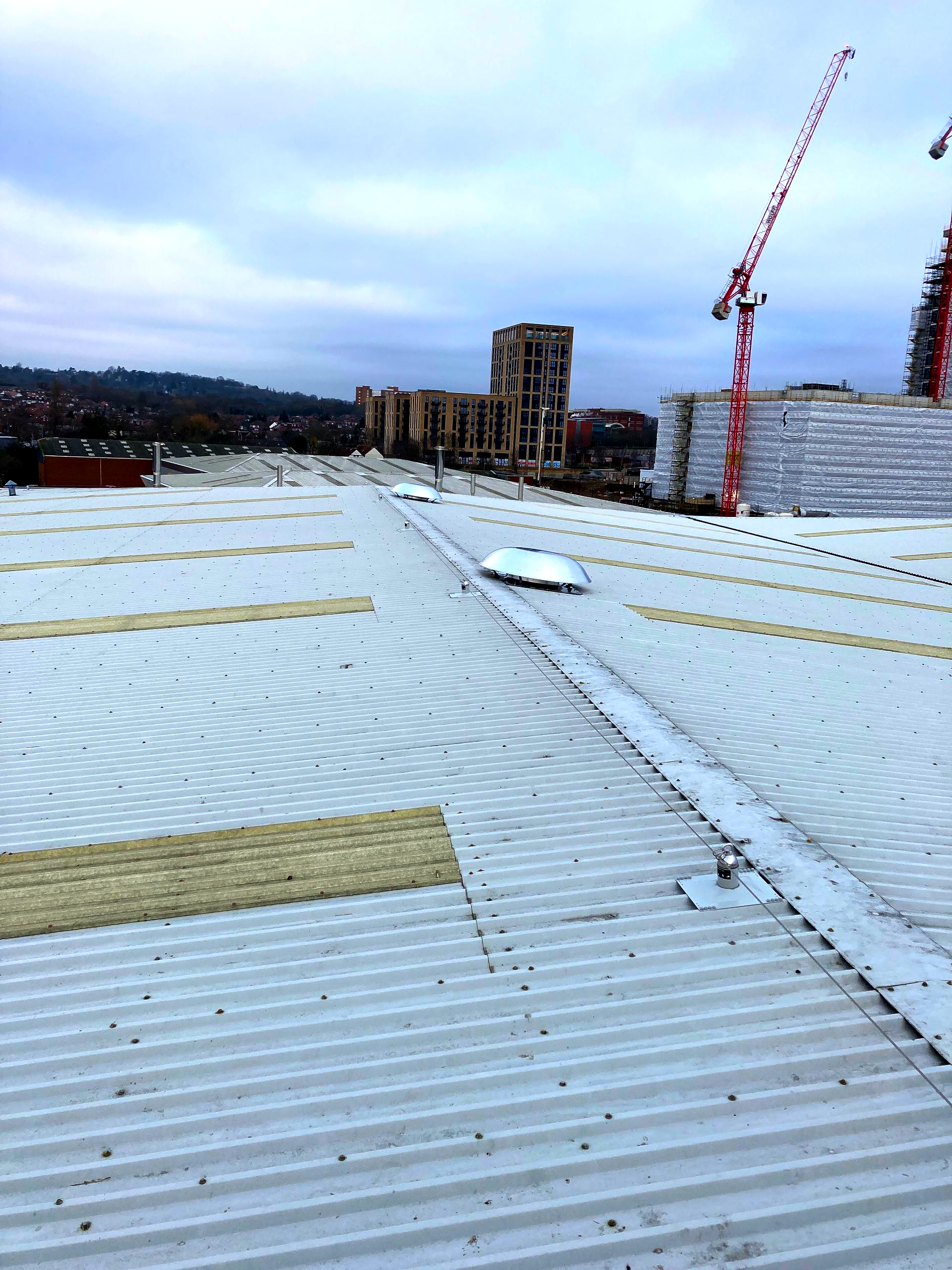 View of a metal roof under construction with insulation strips and a tall crane in the background under a cloudy sky.