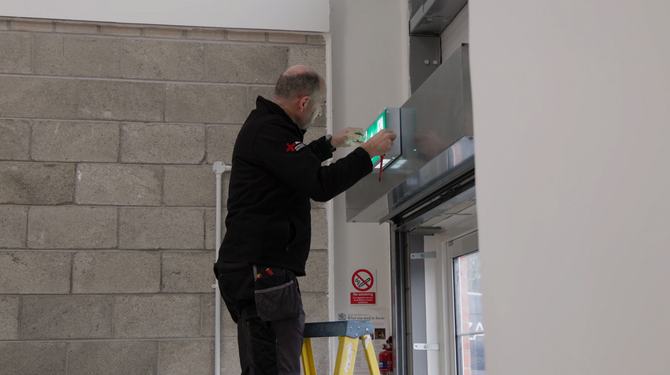 A technician in a high-visibility jacket and hard hat uses a tablet while inspecting electrical switchgear panels.