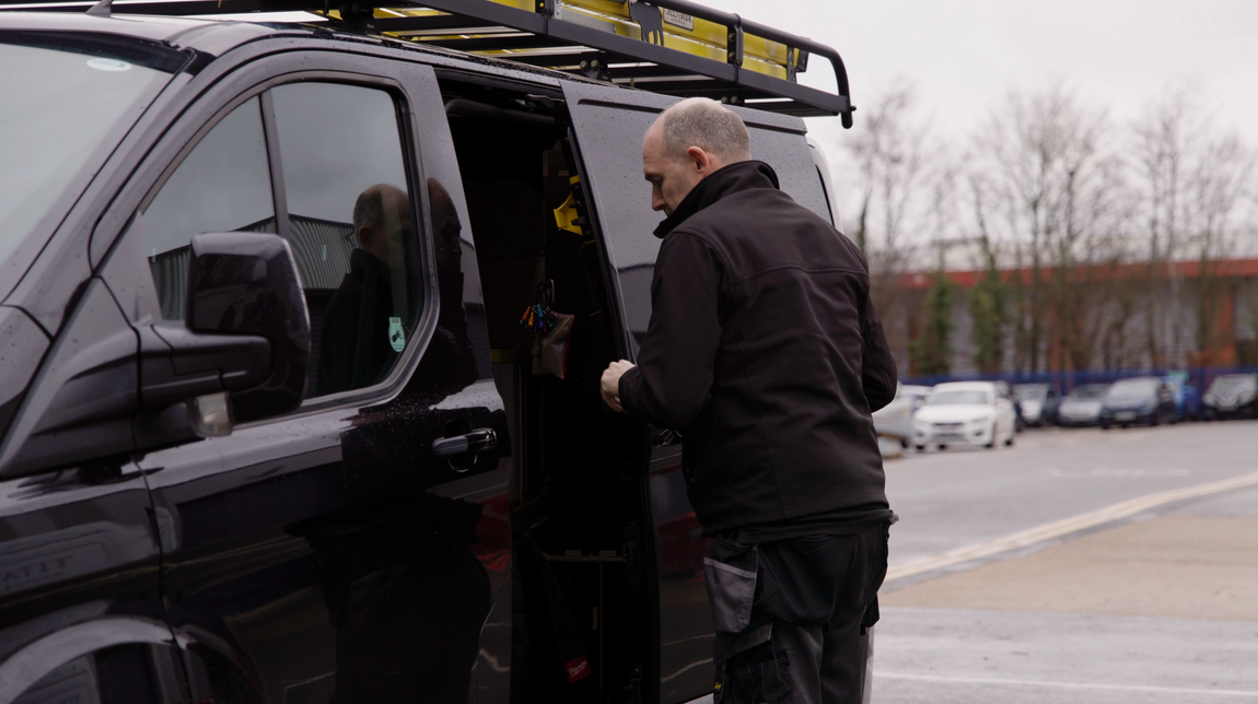 A person in a black jacket and work trousers stands by the open sliding door of a black van in an outdoor parking lot.