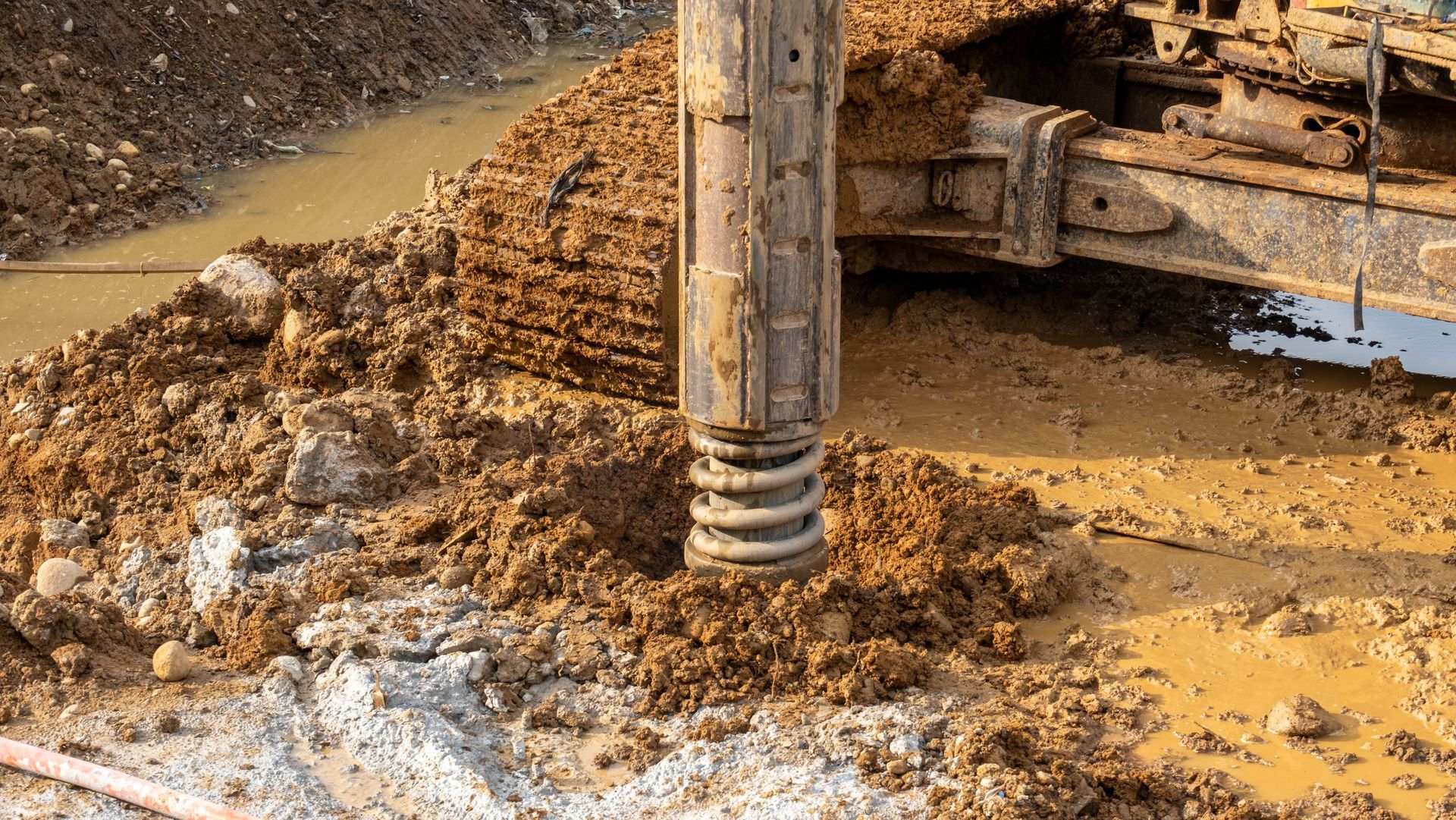 Construction machine drill digging in muddy ground, with wet soil and gravel around the auger.