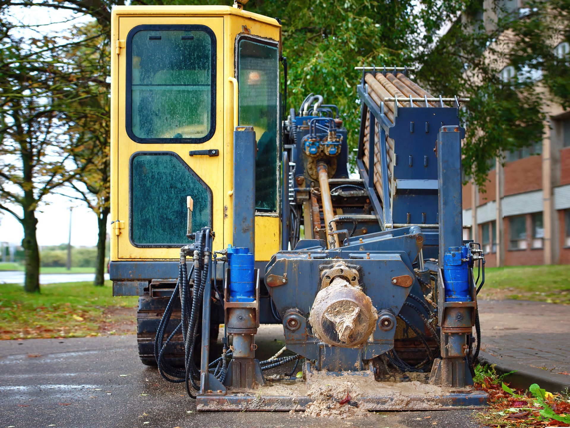 Blue and yellow utility machine with front equipment parked on a wet road beside a building and trees
