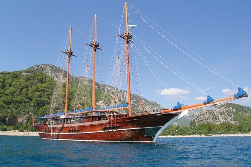 Houten zeilschip op blauw water, nabij een groene, bergachtige kustlijn.