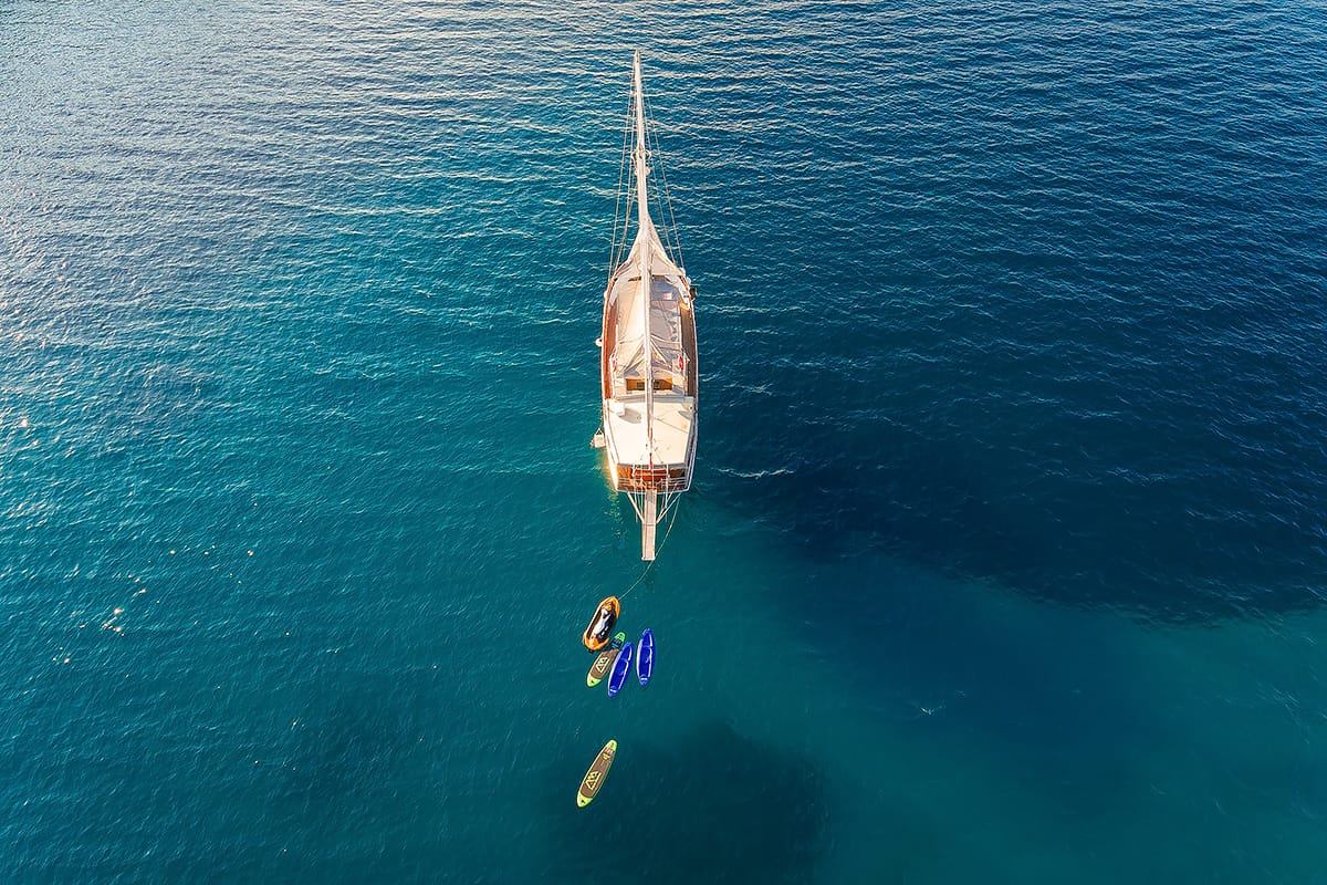 Een houten zeilboot op blauw water, mensen eromheen en twee kajaks.