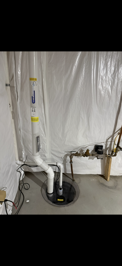 Utility pipes and a vertical white PVC vent in a basement wall, with a floor drain below.