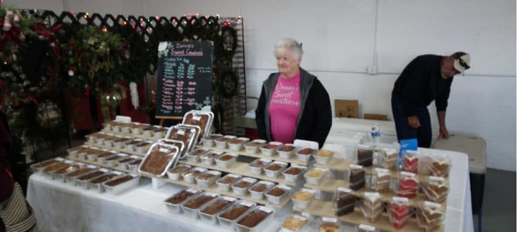 Table of baked goods at a market stall with two vendors behind it and a holiday wreath backdrop