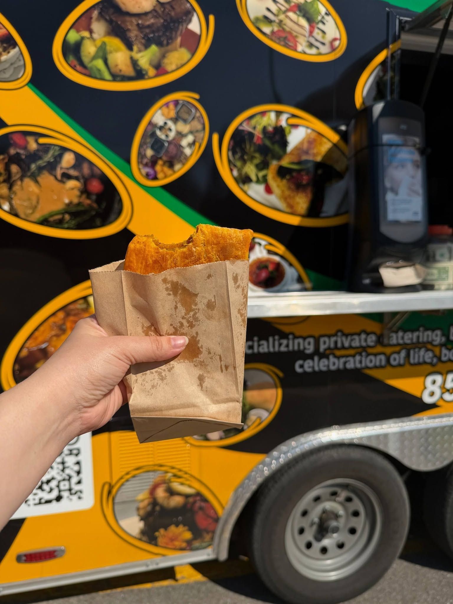 Hand holding a paper bag of fried food in front of a yellow food truck covered with dish photos.