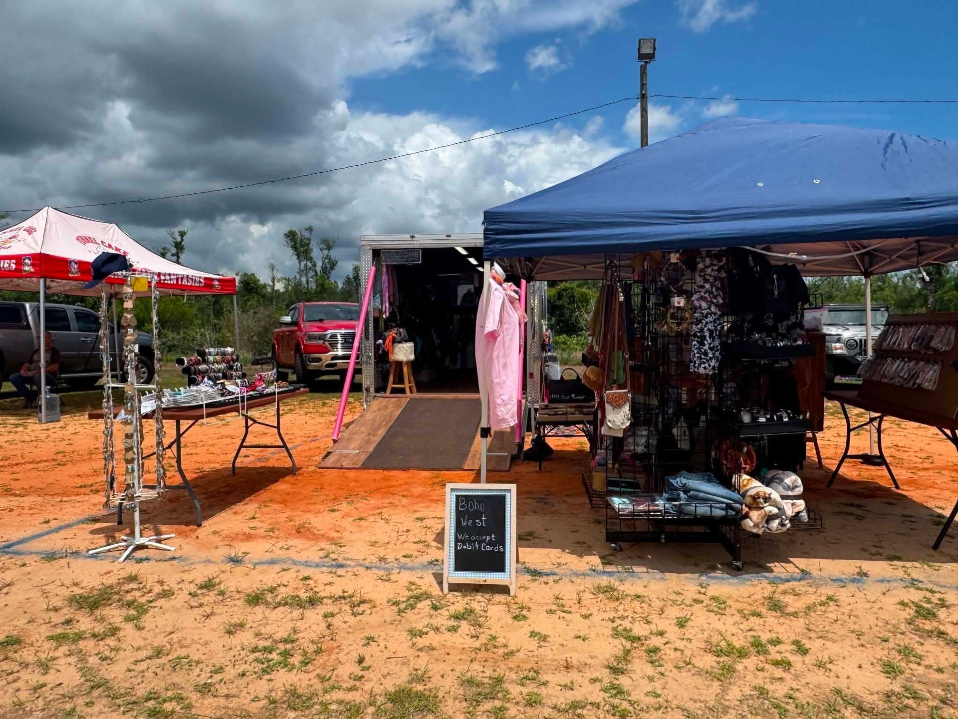 Outdoor market stalls on a cloudy day, with blue and red canopies and clothing racks on orange dirt.