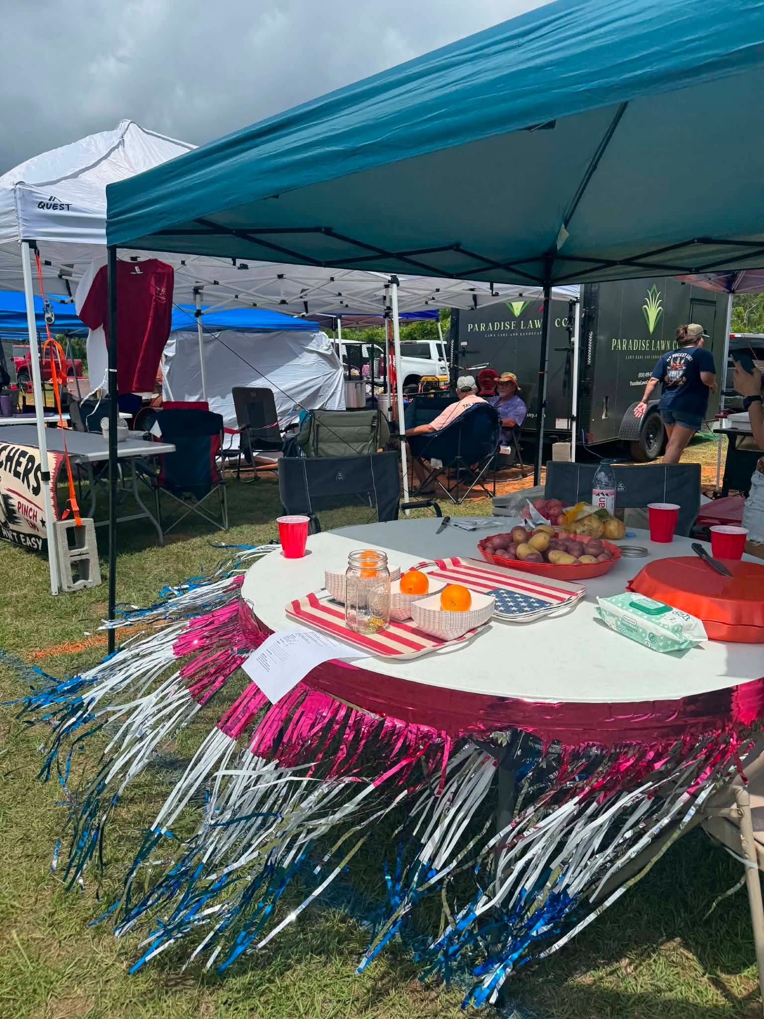 Outdoor party under a blue canopy with a decorated table, food, drinks, and people seated in the shade