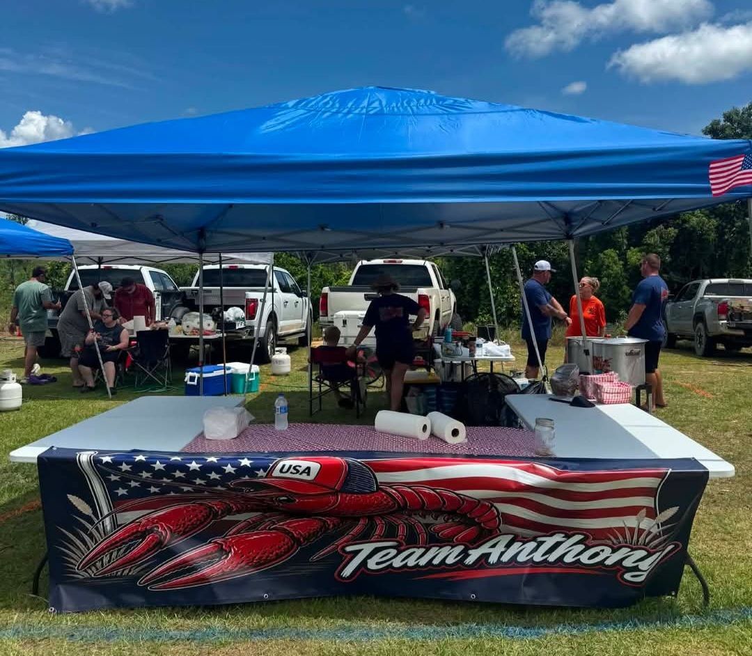 Blue canopy booth with Team Anthony banner at an outdoor event, people and vehicles in the background
