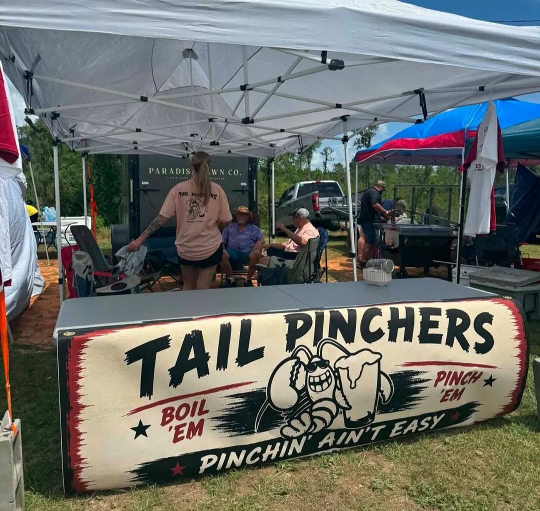 Tail Pinchers food stand under a white tent, with a large hand-painted sign and two people behind the counter