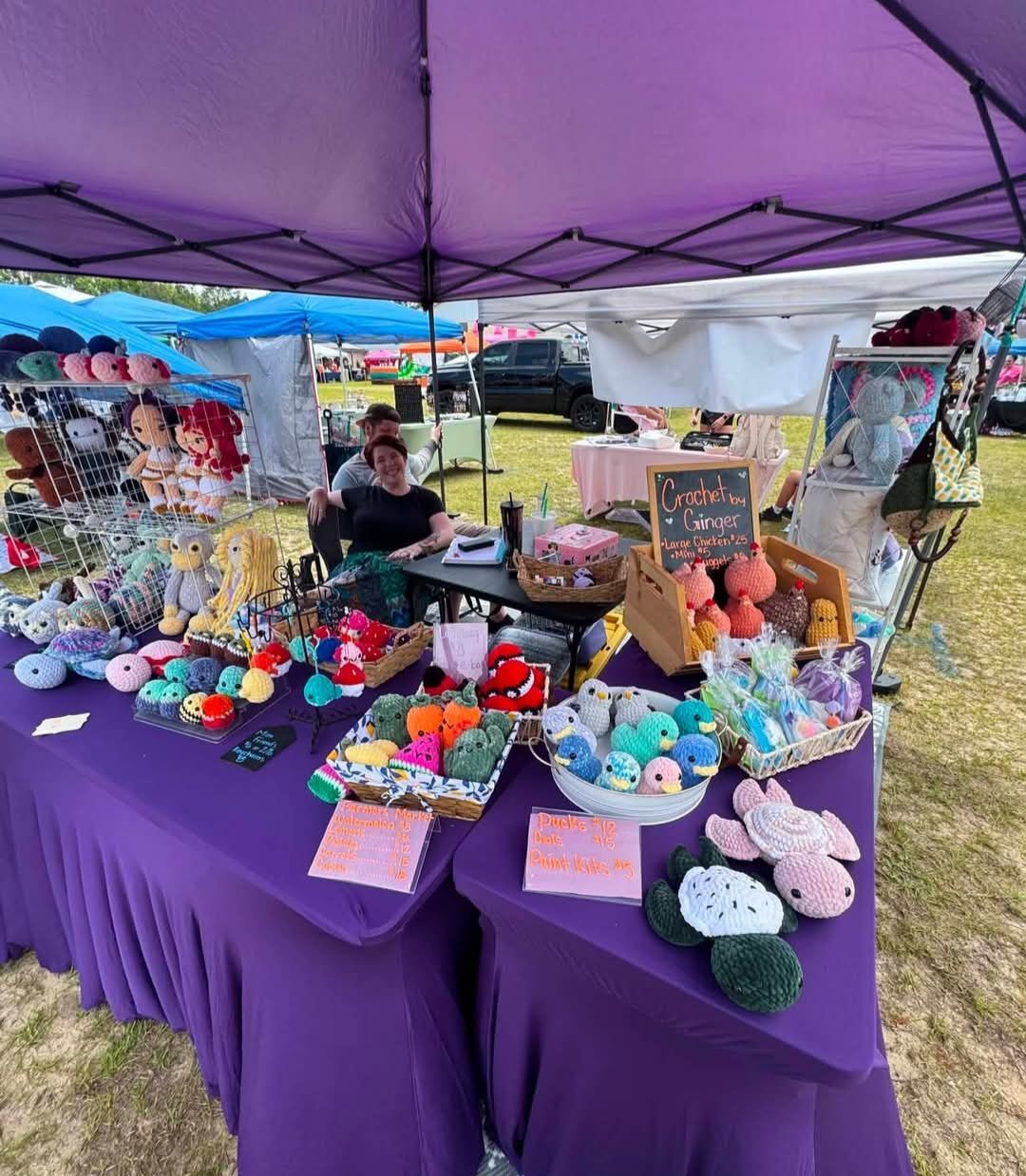 Colorful handmade plush toys and crafts displayed on a purple-covered market stall under a canopy.