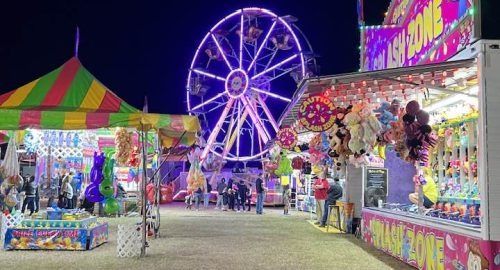 Colorful fairground at night with a lit Ferris wheel and bright carnival stalls along a gravel path