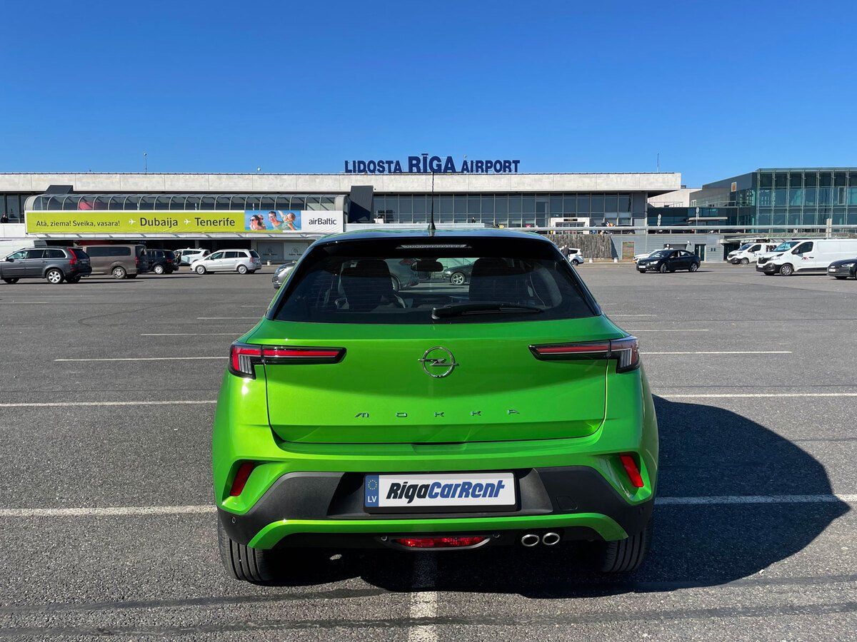 Zaļa automašīna ir novietota stāvvietā lidostas priekšā.