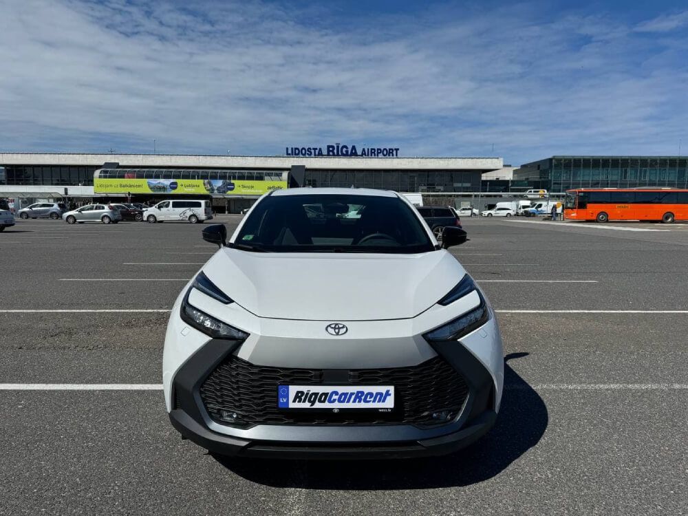 Balta automašīna ir novietota stāvvietā pie Rīgas lidostas