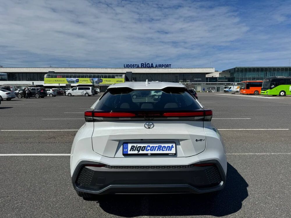 Balta automašīna ir novietota stāvvietā lidostas priekšā.