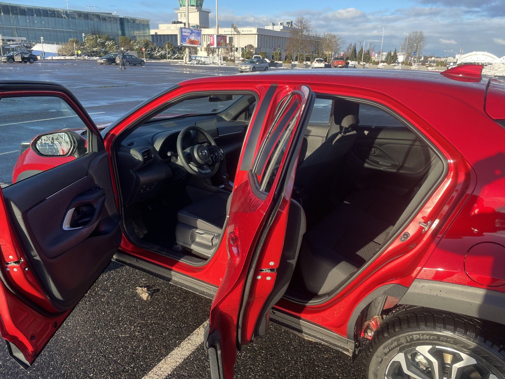 Autostāvvietā ir novietota sarkana automašīna ar atvērtām durvīm.