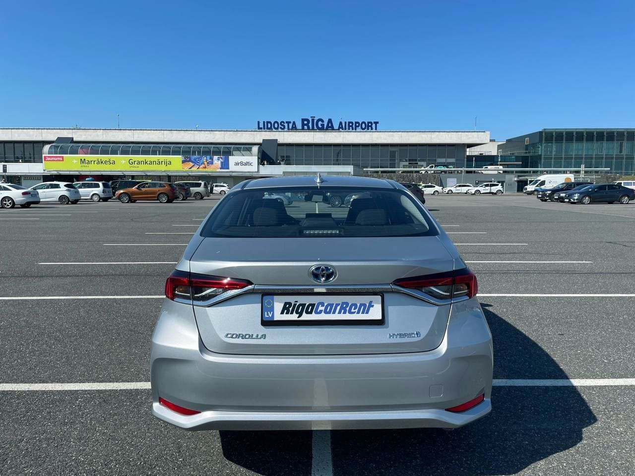 Sudrabaina automašīna ir novietota stāvvietā lidostas priekšā.