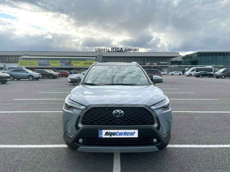 Toyota Corolla Cross ir novietota autostāvvietā lidostas priekšā.