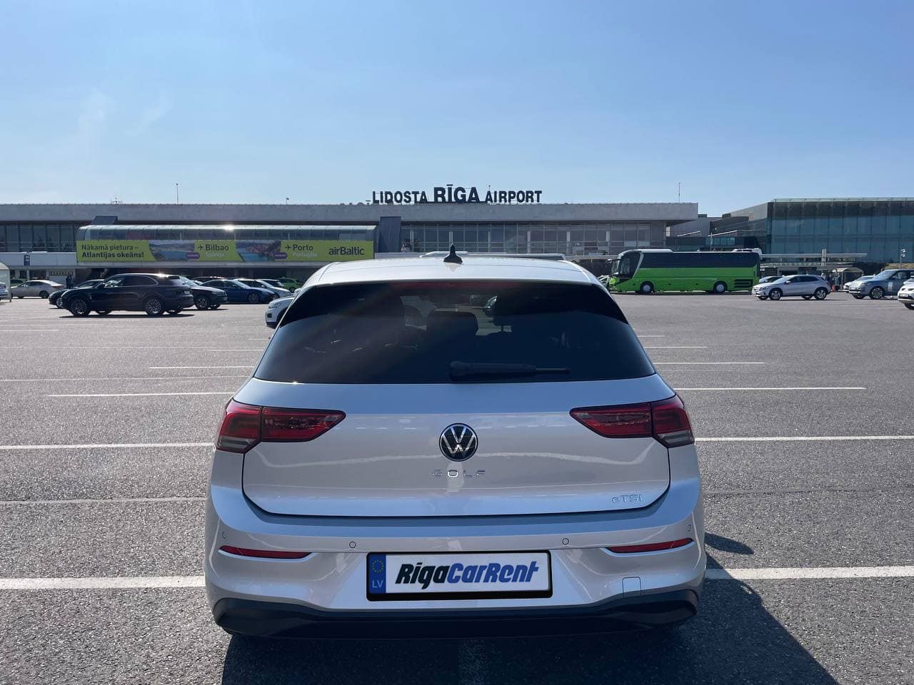 Balta automašīna ir novietota stāvvietā lidostas priekšā.