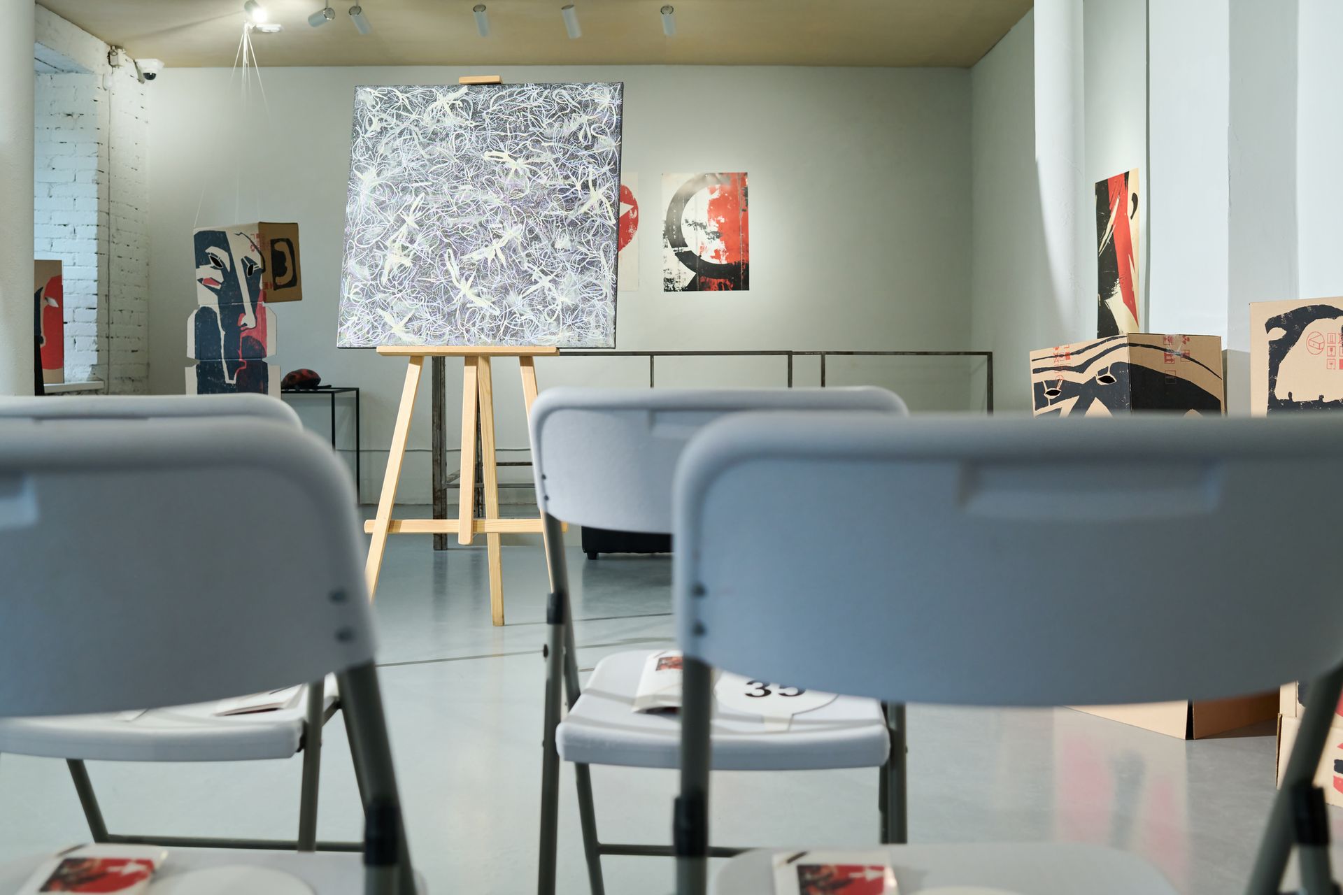 Easel with artwork standing in the center of a fine art gallery in front of white plastic chairs.