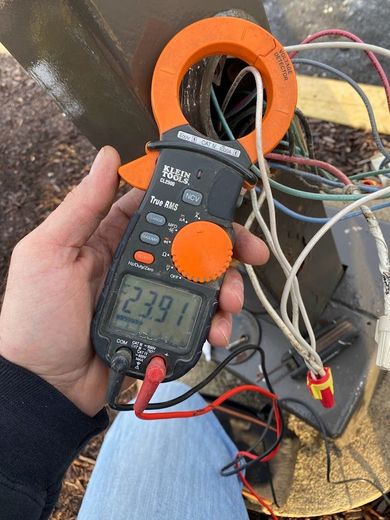 A hand holding a multimeter measuring amperage on an outdoor HVAC unit; reads 23.91.