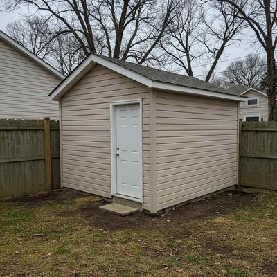 Small beige backyard shed with white door, surrounded by a wooden fence and leafless trees.