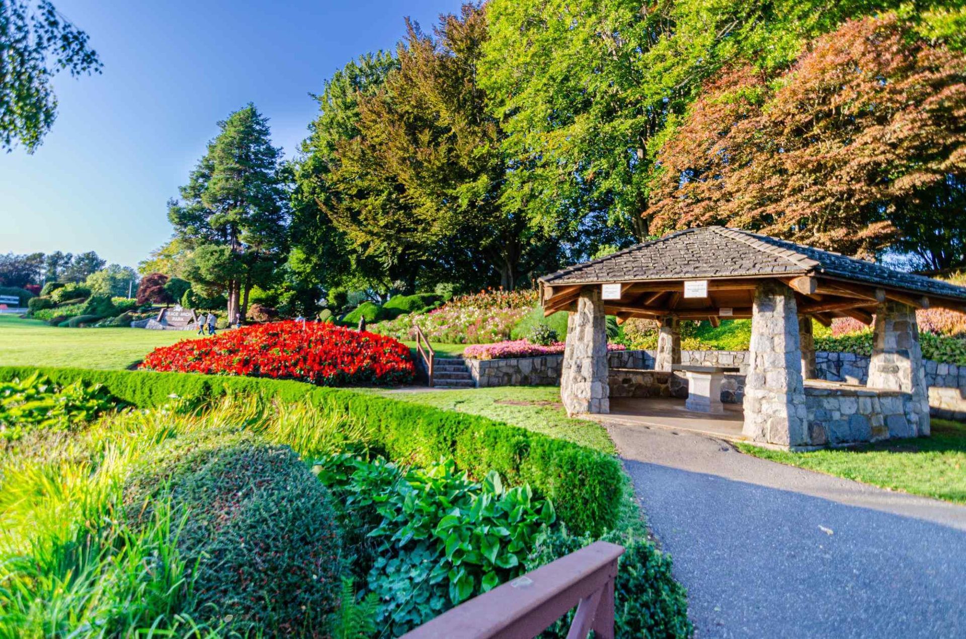 Sunny park with colorful flower beds, green lawn, and a stone gazebo beside a paved path