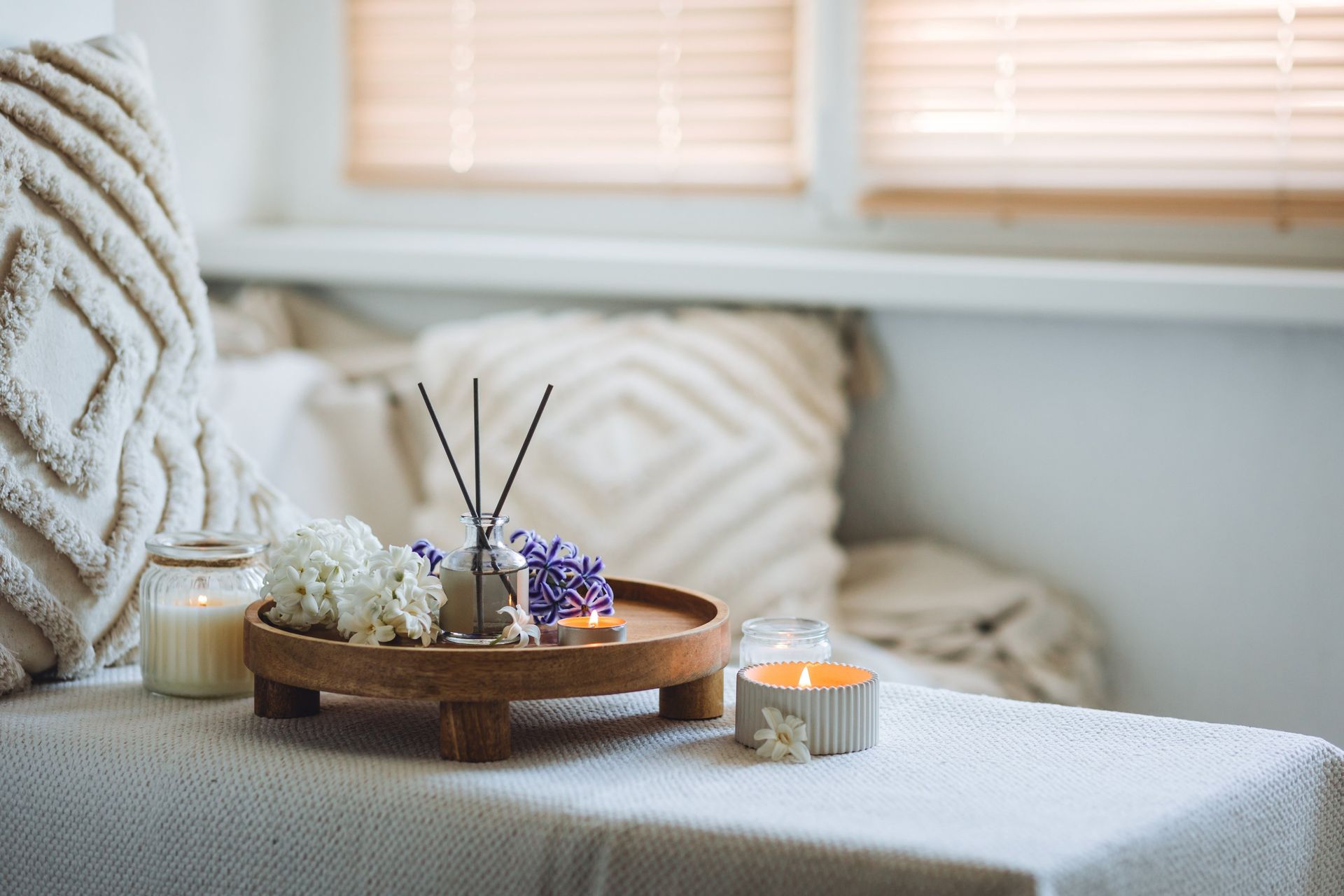 Cozy white sofa with textured pillows, wooden tray, candles, flowers, and reed diffuser by a sunlit window