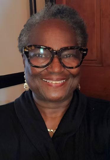 Smiling person wearing large black glasses and a black top, standing indoors against a wooden cabinet.