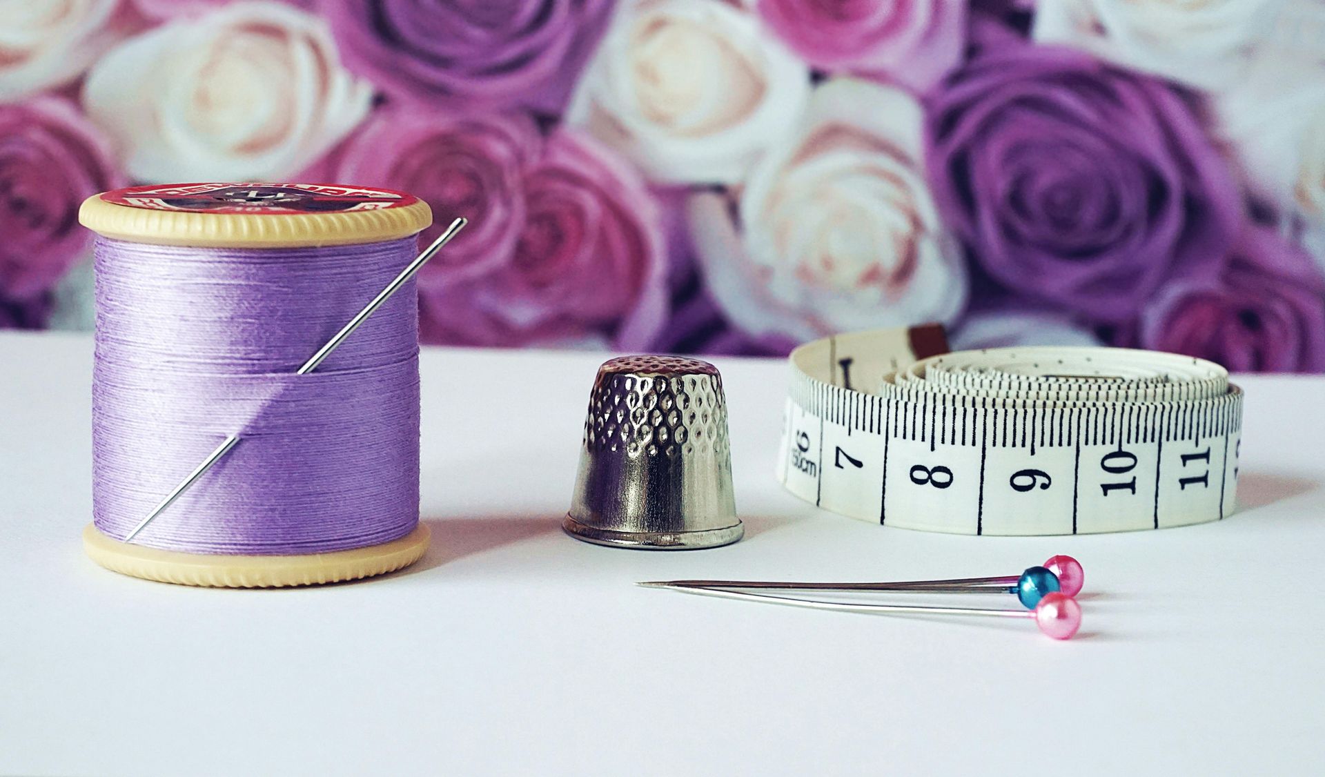 A spool of purple thread, a thimble, a measuring tape, and sewing pins arranged on a white surface before blurred roses.