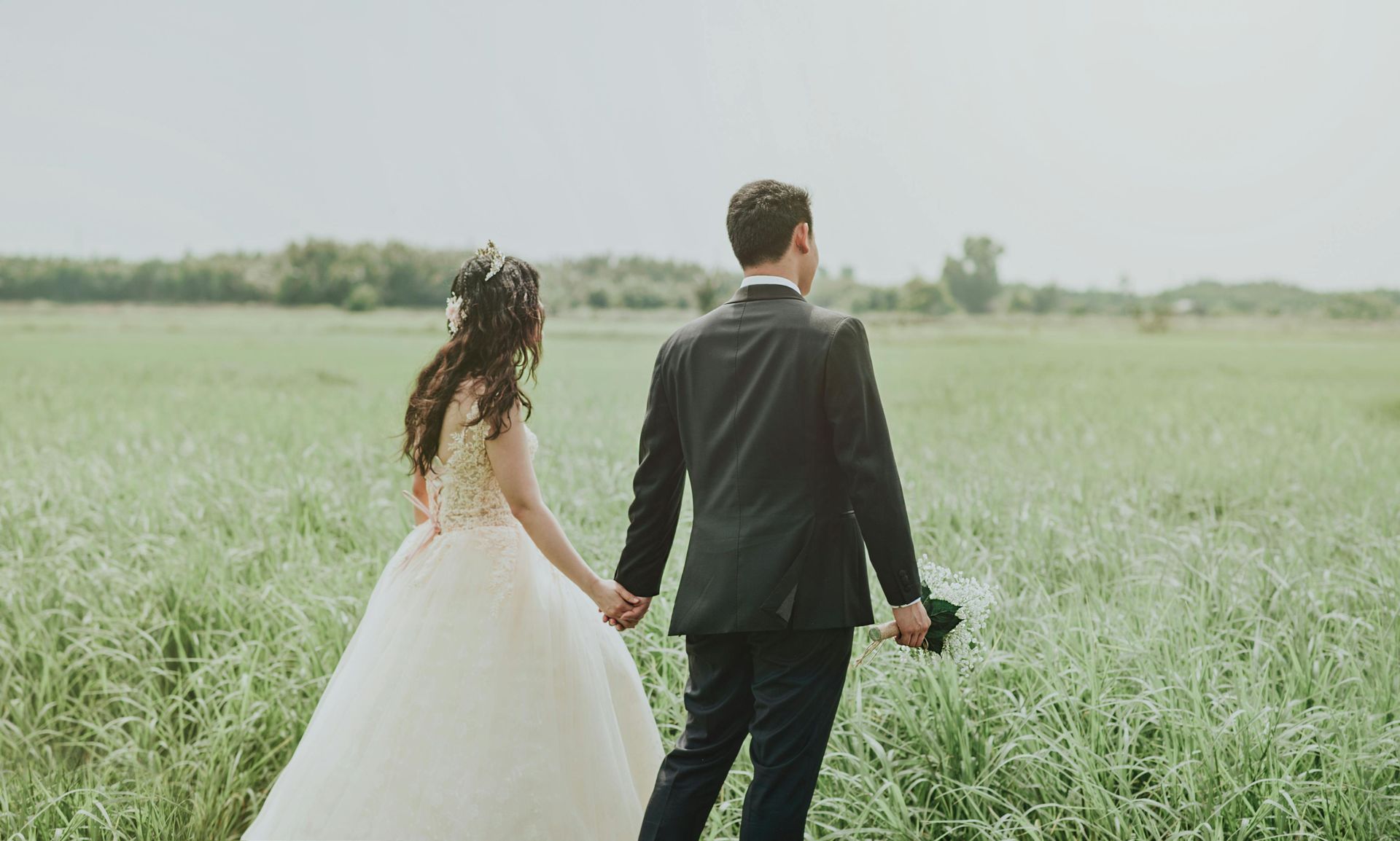 A couple in formal wedding attire walks hand-in-hand through a grassy field.