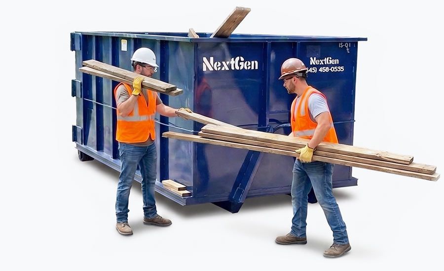 Two workers in orange vests and hard hats carry long metal rods beside a blue dumpster.