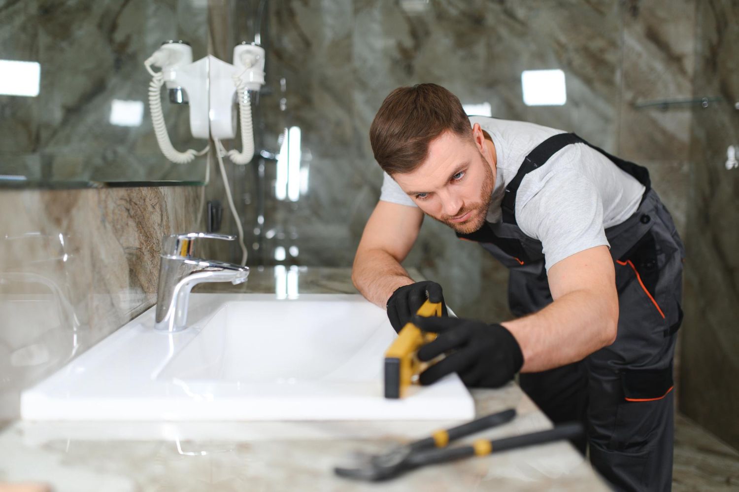 A professional in work coveralls uses a spirit level to check the alignment of a bathroom sink on a marble countertop.