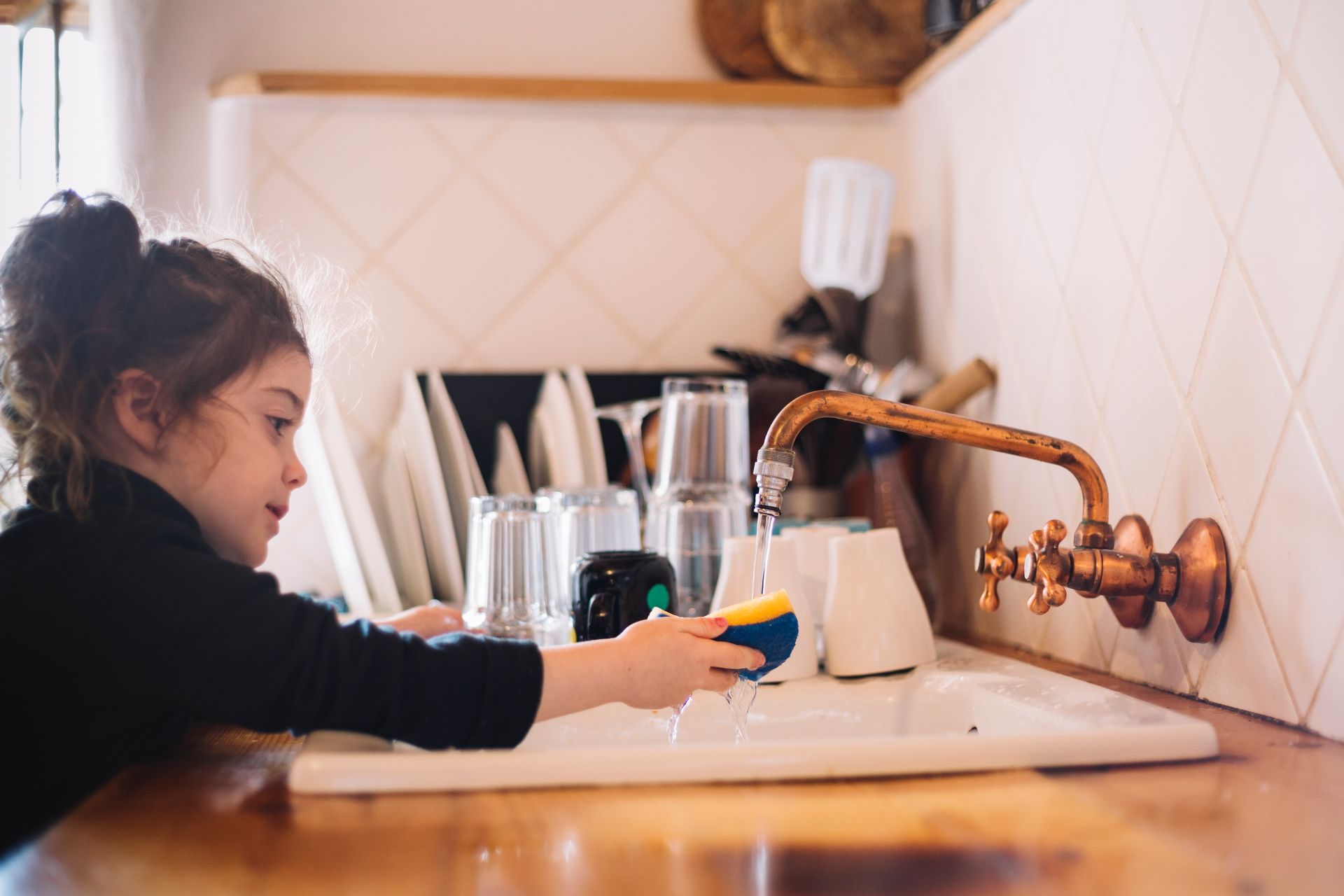 A little girl is washing dishes in a kitchen sink.