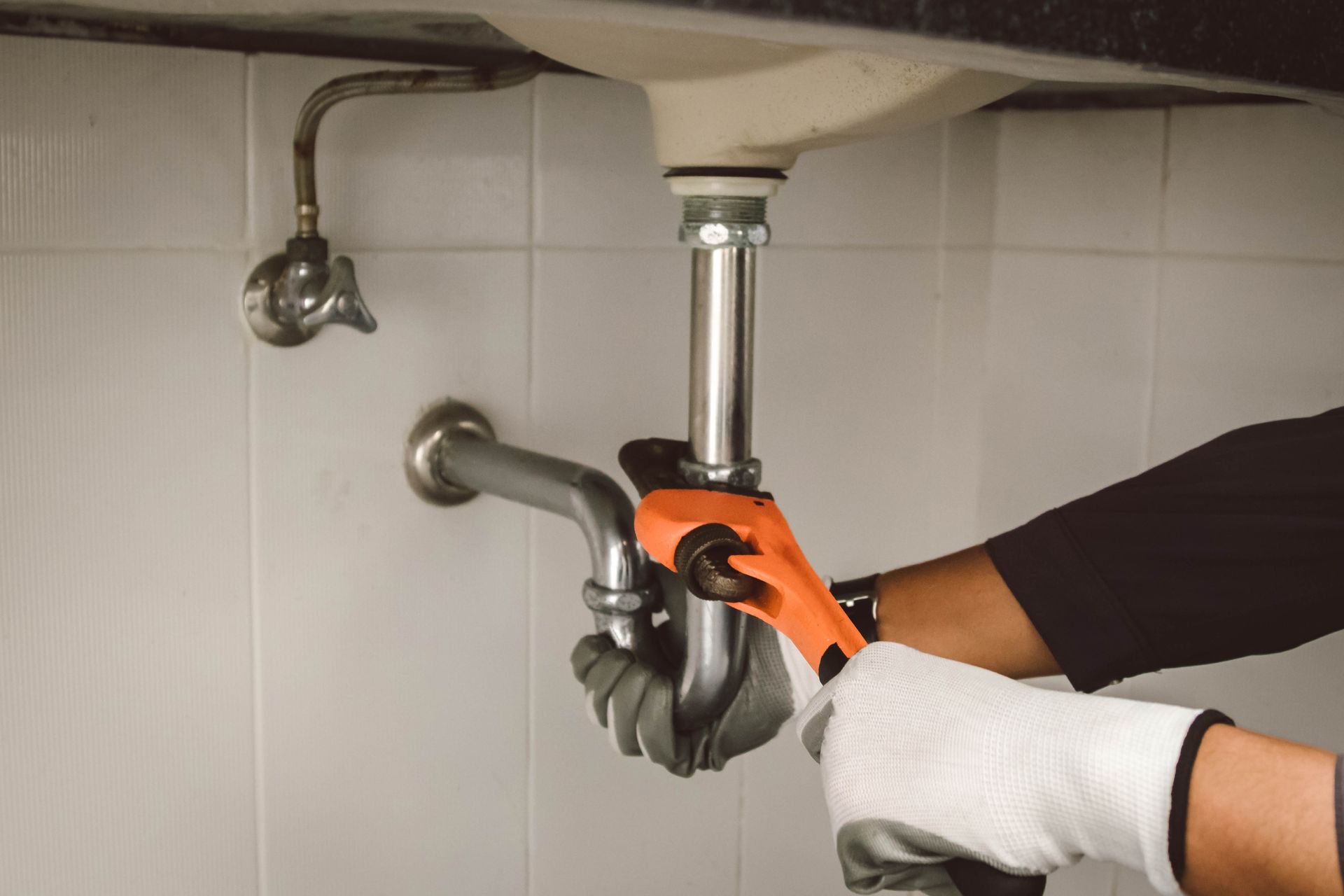 Person using an orange wrench to tighten plumbing under a sink in a tiled bathroom.