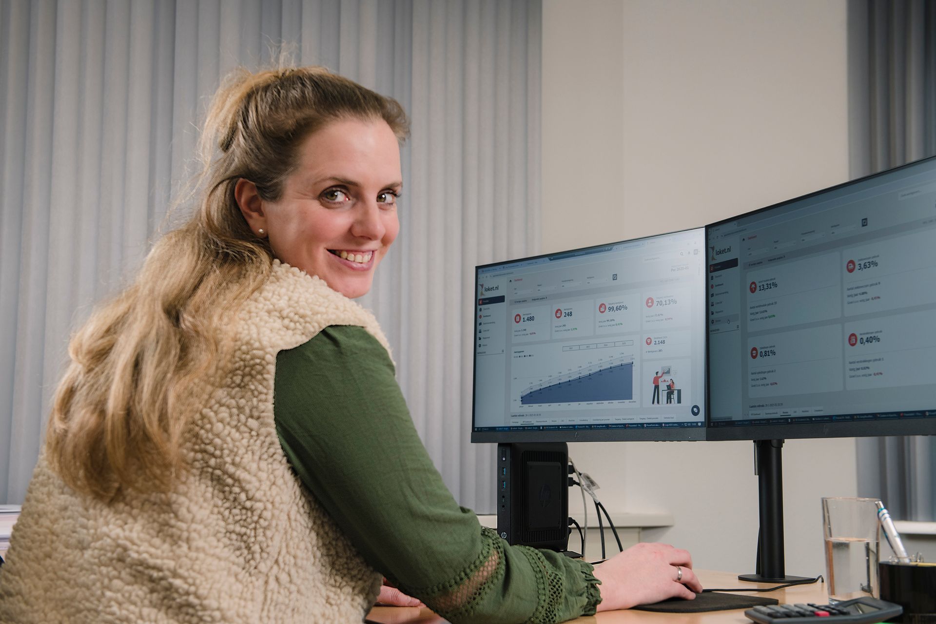 Een vrouw glimlacht naar twee beeldschermen in een kantoor en bekijkt grafieken op de schermen.