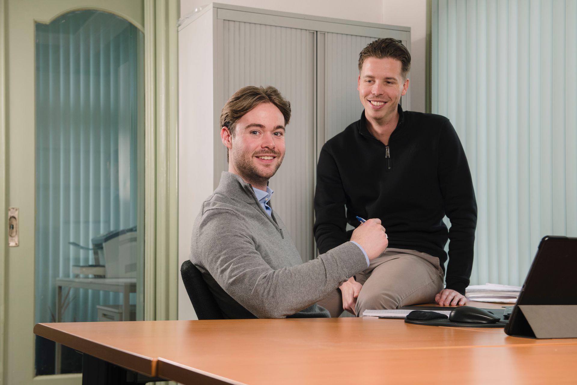 Twee mannen in een vergaderruimte, zittend aan een bureau met een laptop en lachend naar de camera.