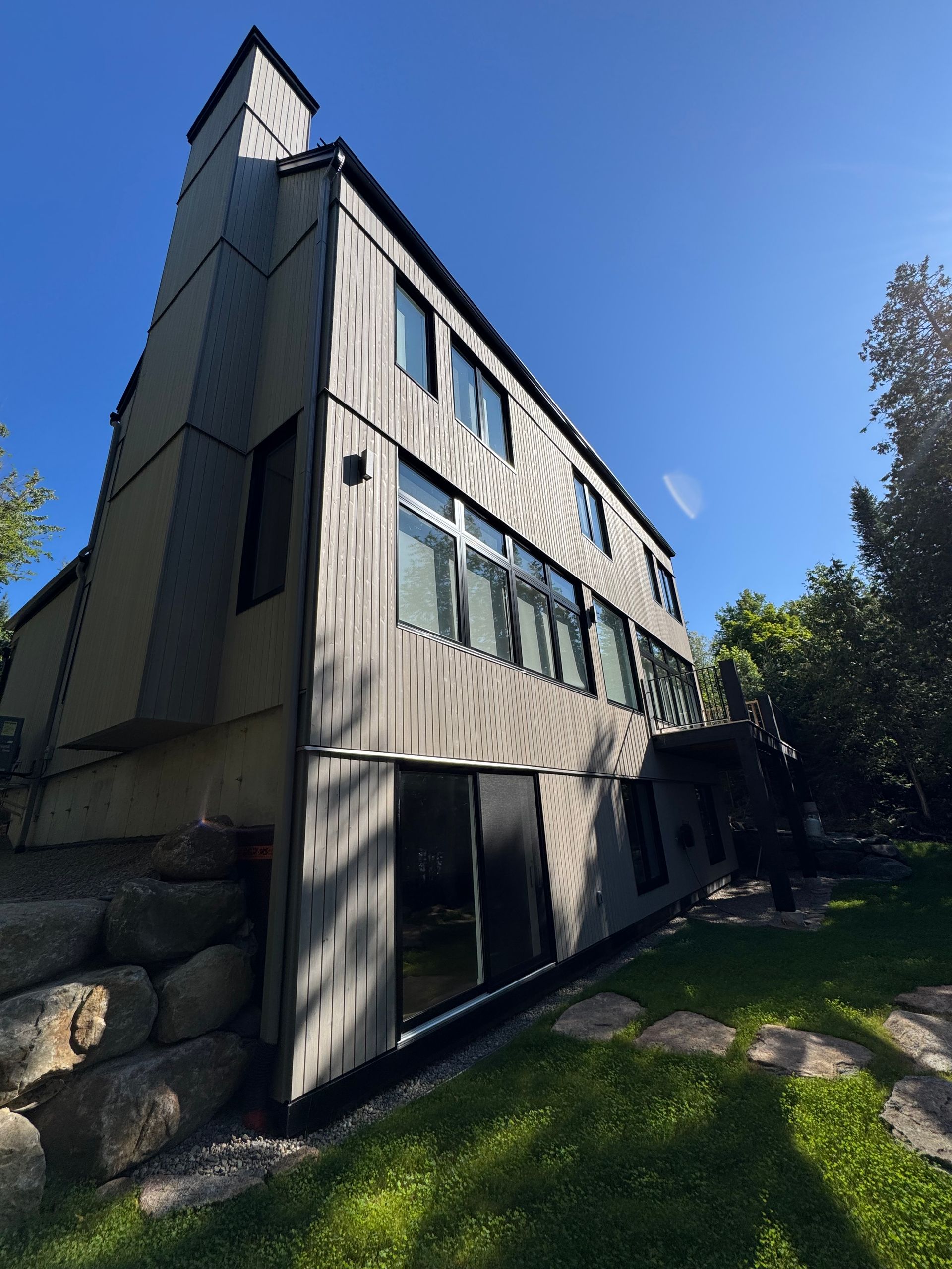 Maison grise moderne avec de grandes fenêtres, une haute cheminée et un jardin gazonné sous un ciel bleu.