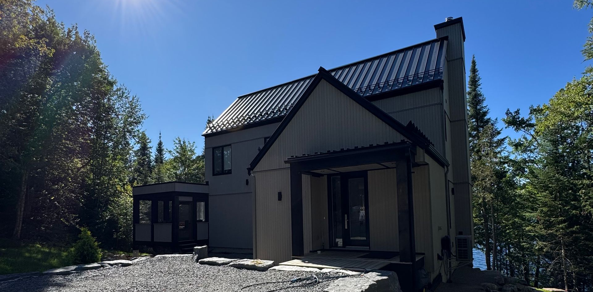 Maison grise moderne avec toit noir, entourée d'arbres et d'un ciel bleu azur.
