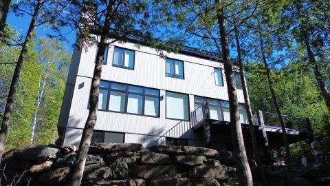 Maison moderne de trois étages avec un revêtement gris, nichée dans une zone boisée, entourée d'arbres et d'un mur de pierre.