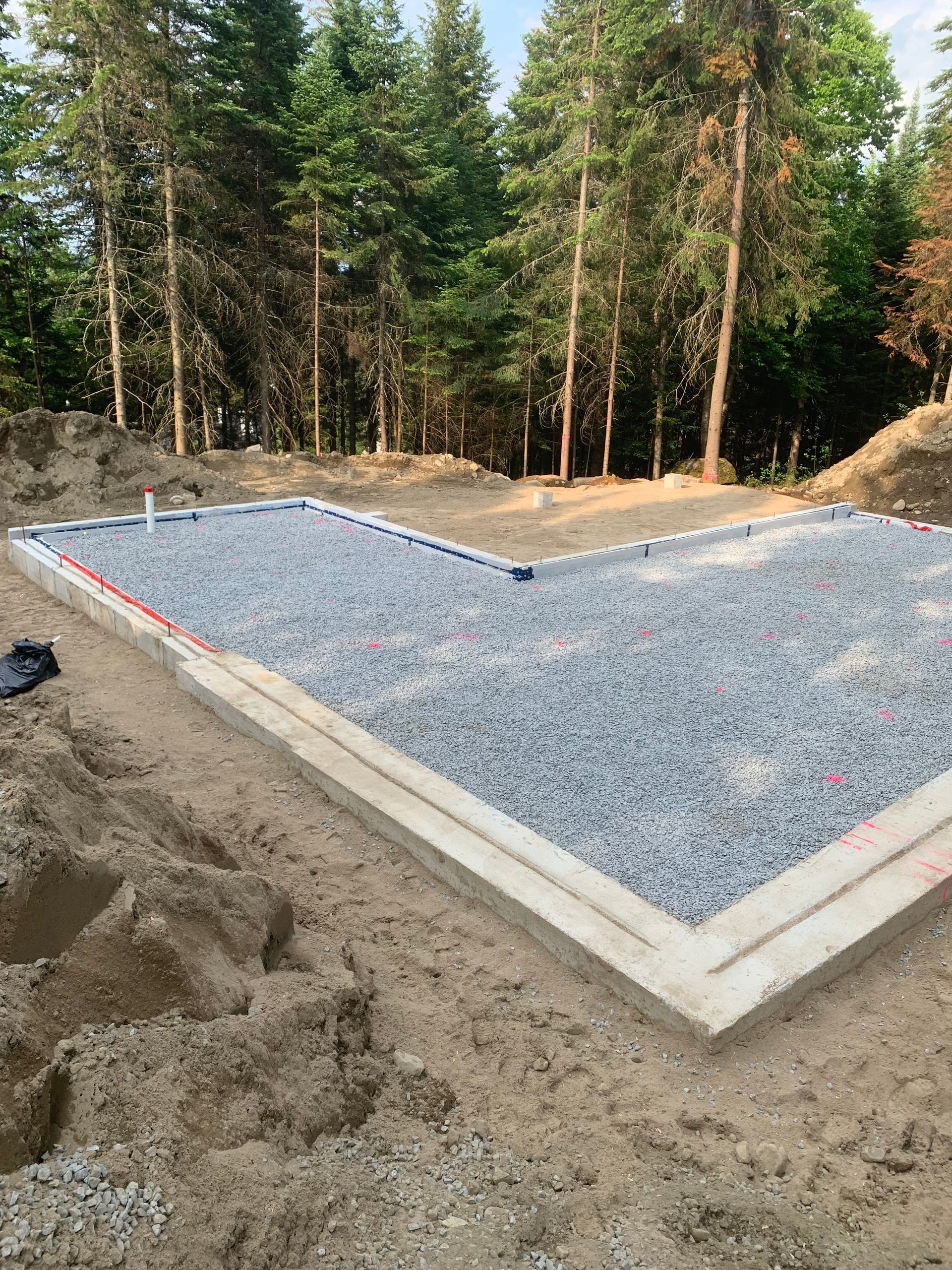 Fondation en béton en forme de L remplie de gravier, en attente de construction dans une zone boisée.