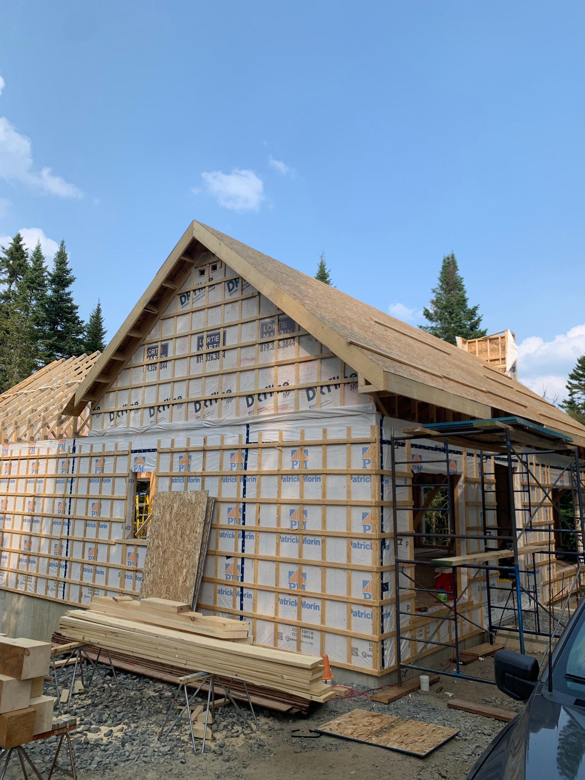 Maison en construction, vue extérieure. Murs recouverts d'isolant et de bois, toit partiellement couvert de bardeaux. Ciel dégagé et arbres en arrière-plan.