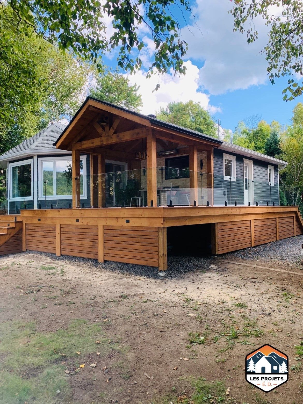 Terrasse en bois avec porche couvert attenant à une maison aux balustrades en verre, entourée d'arbres.