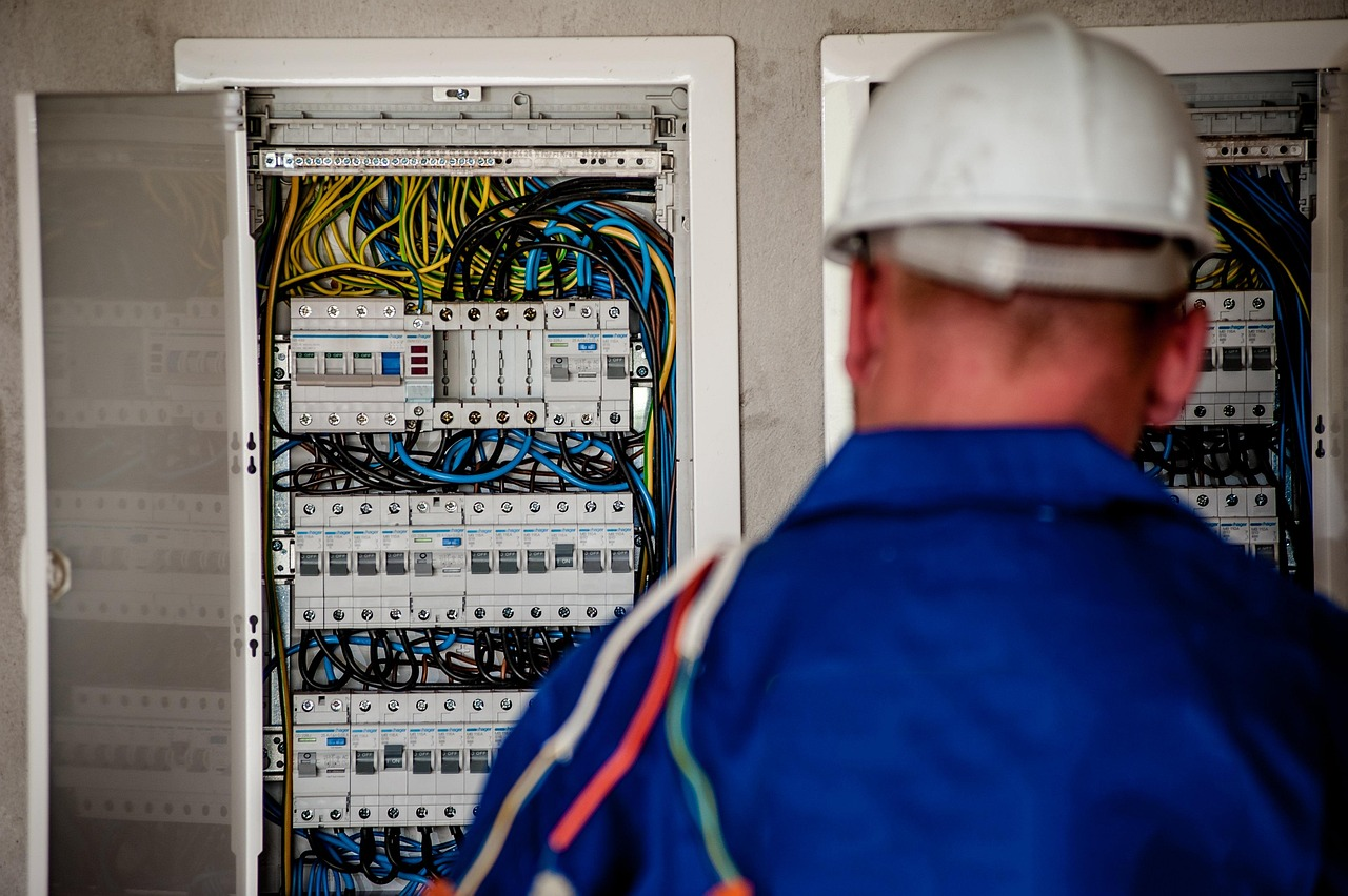 Electrician in blue uniform and white helmet inspects an electrical panel, wires visible.
