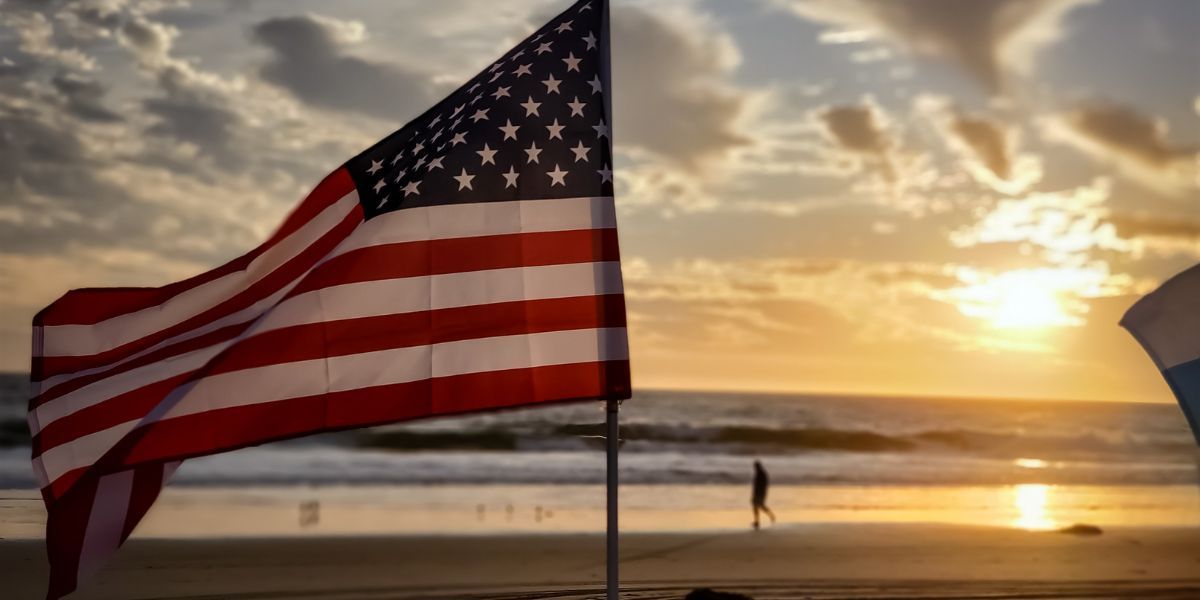 A close-up of the American flag waving in the breeze with a serene sunrise in the background. The sky is filled with soft clouds, and the ocean waves gently lap at the shore, with a silhouette of a lone person walking along the beach.
