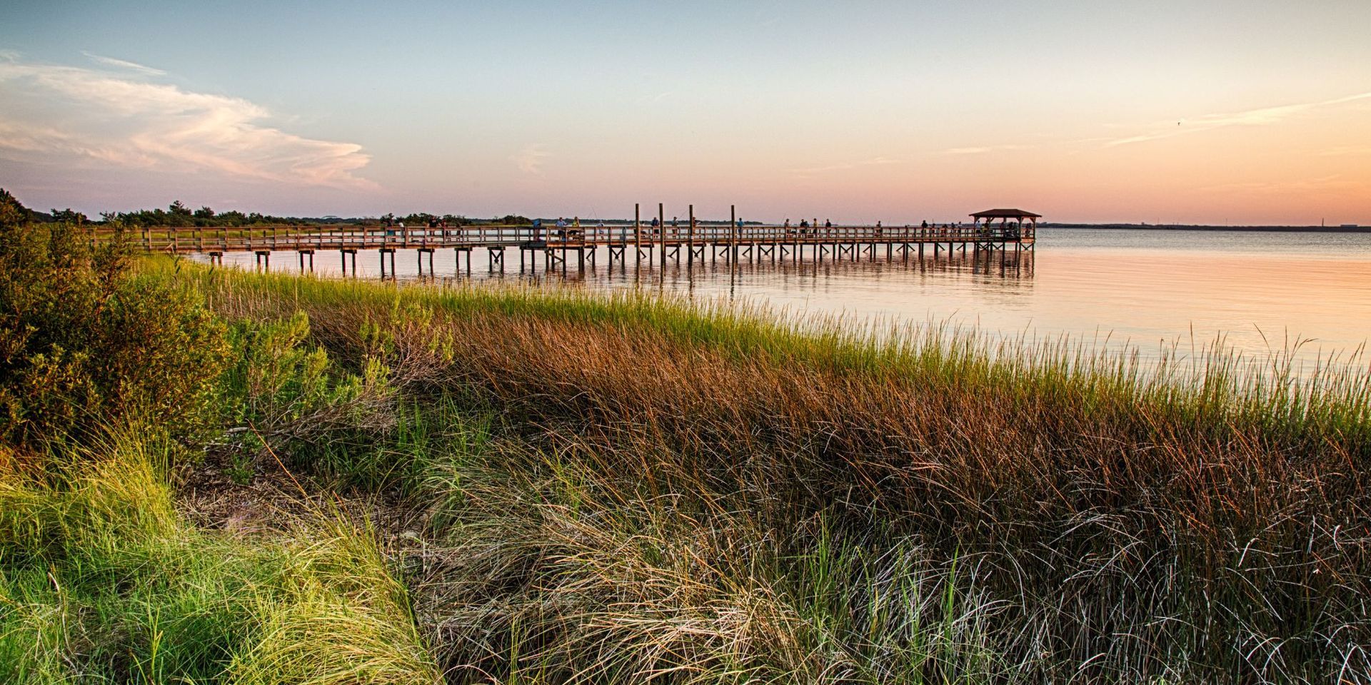 A long wooden pier extending over the calm waters of the Cape Fear River, surrounded by tall grass