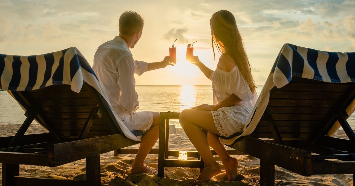 A couple relaxes on wooden lounge chairs at Carolina Beach, toasting with tropical drinks as the sun sets over the ocean. Blue and white striped towels drape over the chairs, and the golden light reflects off the waves, creating a romantic and tranquil atmosphere.