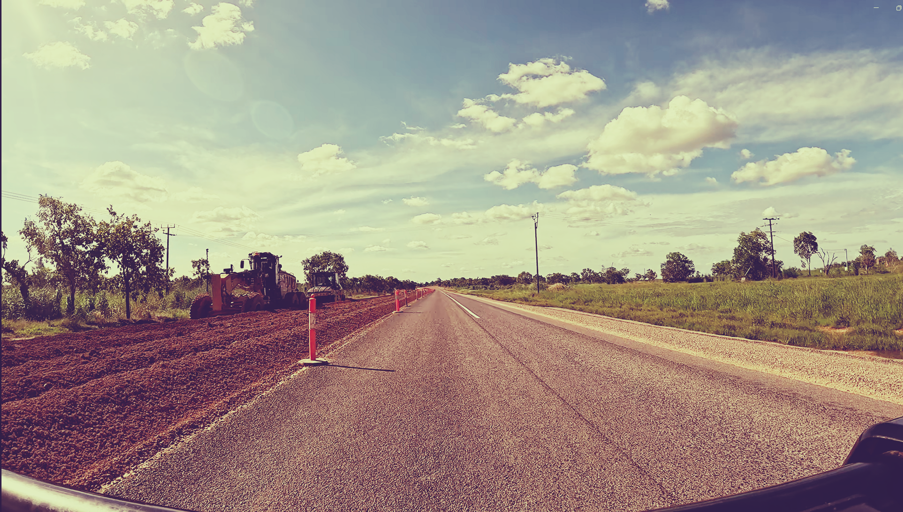 Straight rural road through open fields under a cloudy sky, viewed from a moving vehicle.